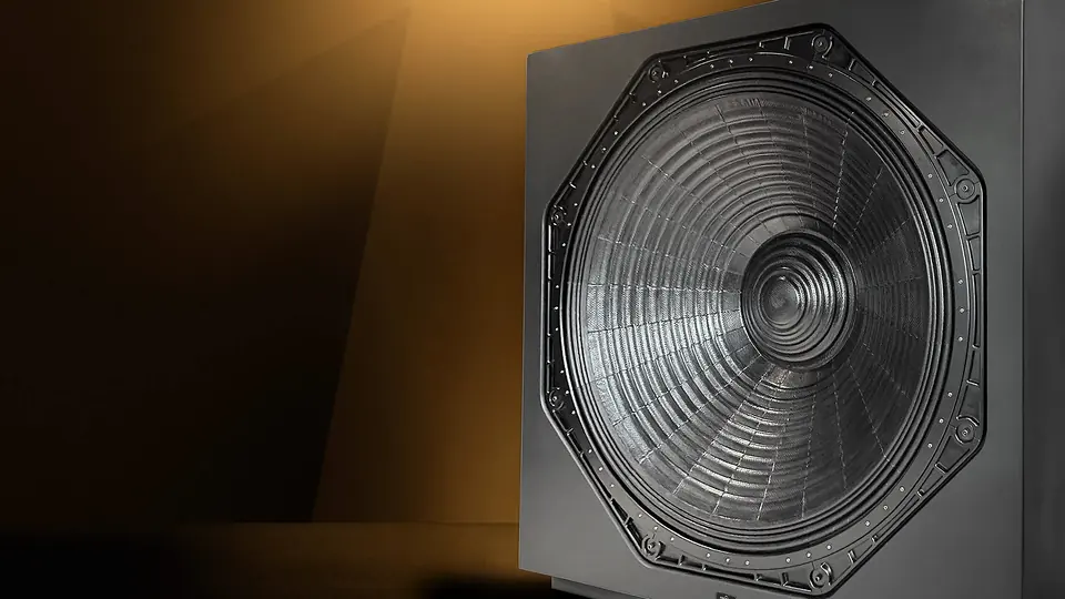 Subwoofer infrasónico Ascendo THE64 SUB Pro en penumbra: su gran cono octagonal de fibra iluminado por un haz de luz dorada desde lo alto.