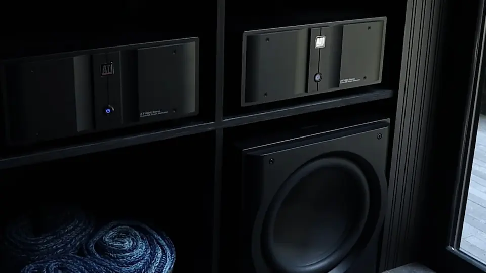 Dos amplificadores ATI negros en un mueble de entrepaños oscuros, con un subwoofer debajo y cable enrollado al pie, en penumbra.
