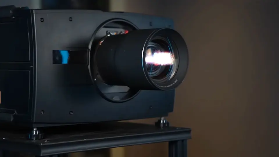 Proyector láser Barco Residential de perfil, con el lente encendido, sobre un soporte en penumbra cálida.