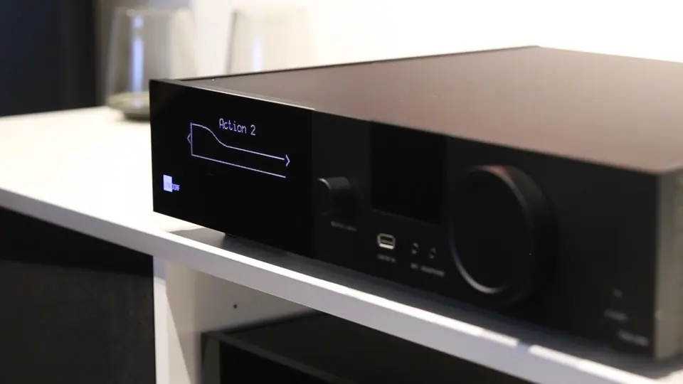 Amplificador integrado Lyngdorf negro sobre una repisa blanca; en su pantalla, la curva de sonido «Action 2».