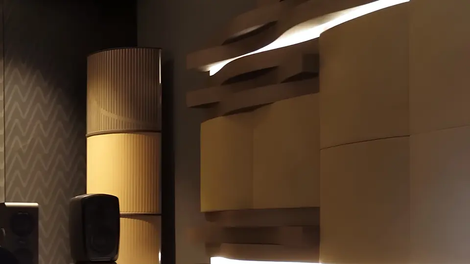 Sala de muestra Univicoustic: columna difusora curva de madera clara y paneles ondulados con luz indirecta, junto a un monitor de estudio.