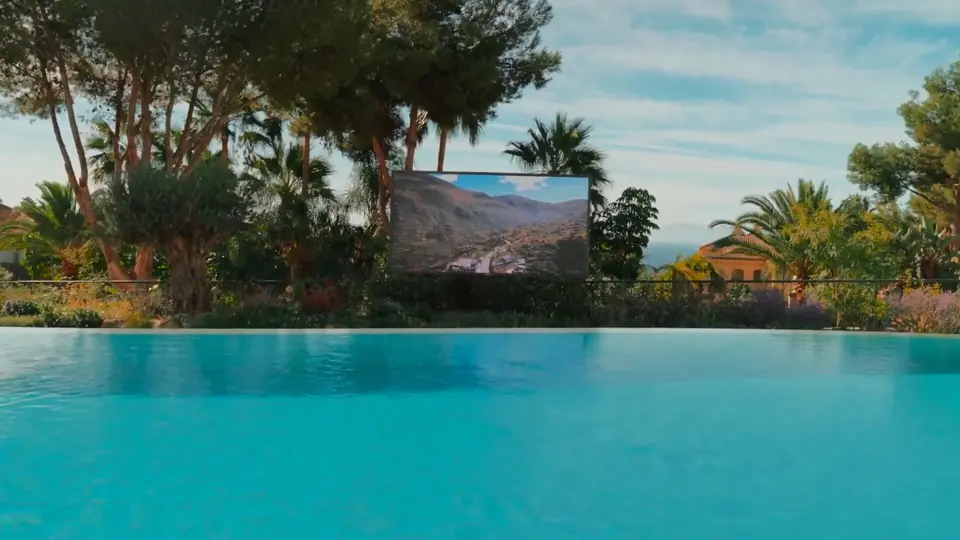 Pantalla exterior Zion entre pinos y palmeras, del otro lado de una alberca infinita, con un paisaje de montaña en la imagen.