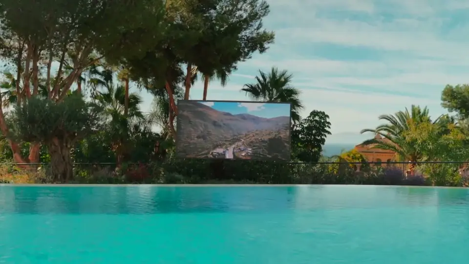Pantalla exterior Zion entre pinos y palmeras, del otro lado de una alberca infinita, con un paisaje de montaña en la imagen.